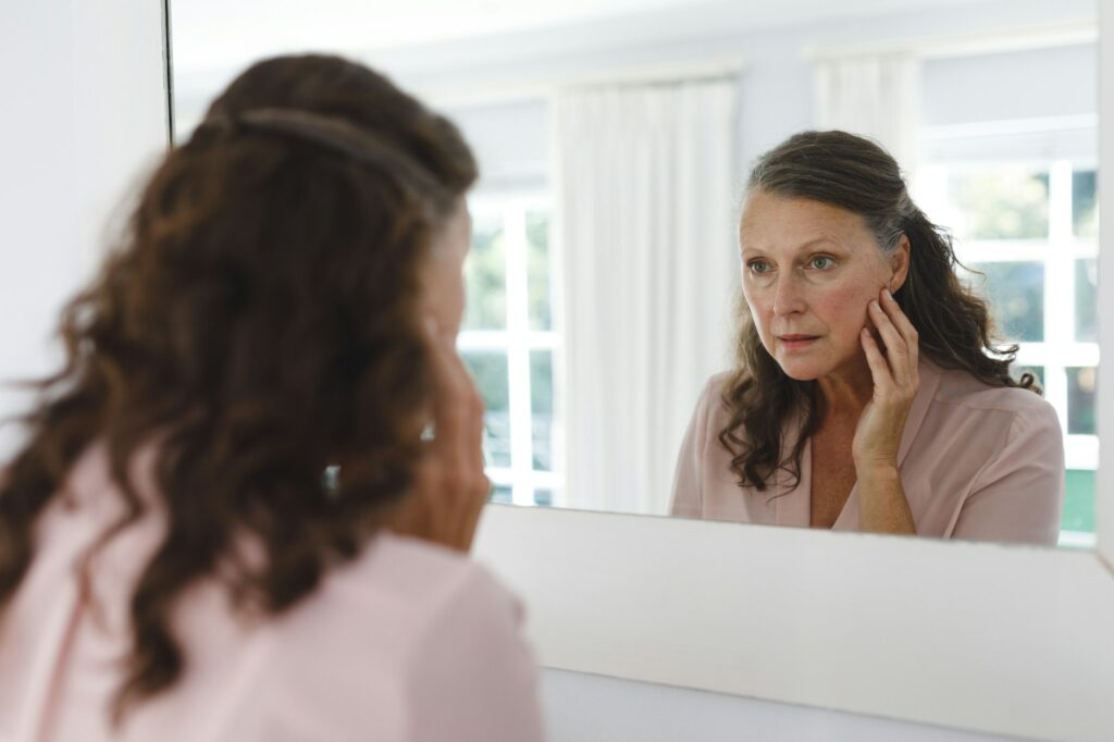 Senior caucasian woman in bathroom, looking at her face in mirror