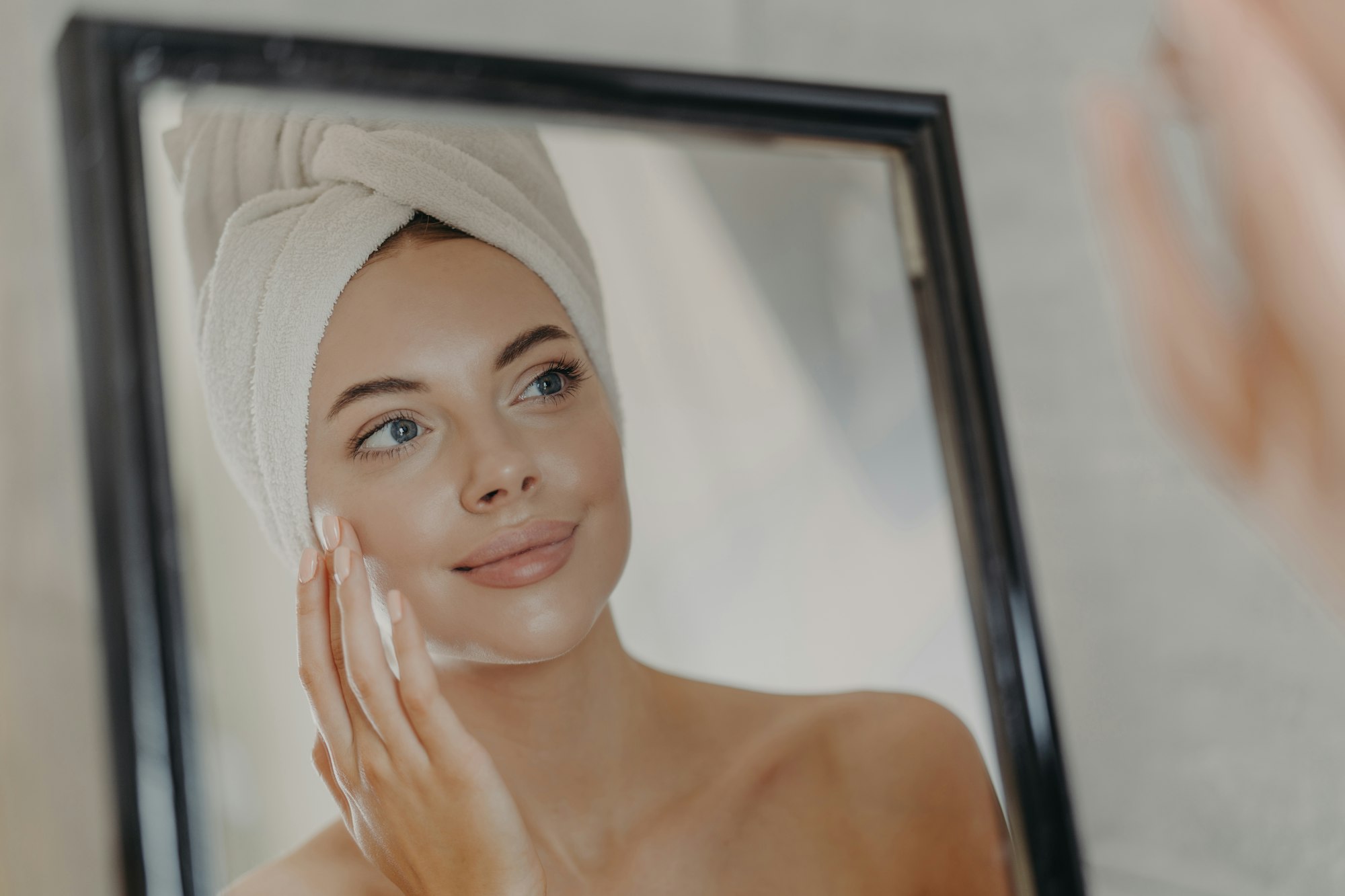 Young naked European woman touches soft skin on face, looks at herself in bathroom mirror